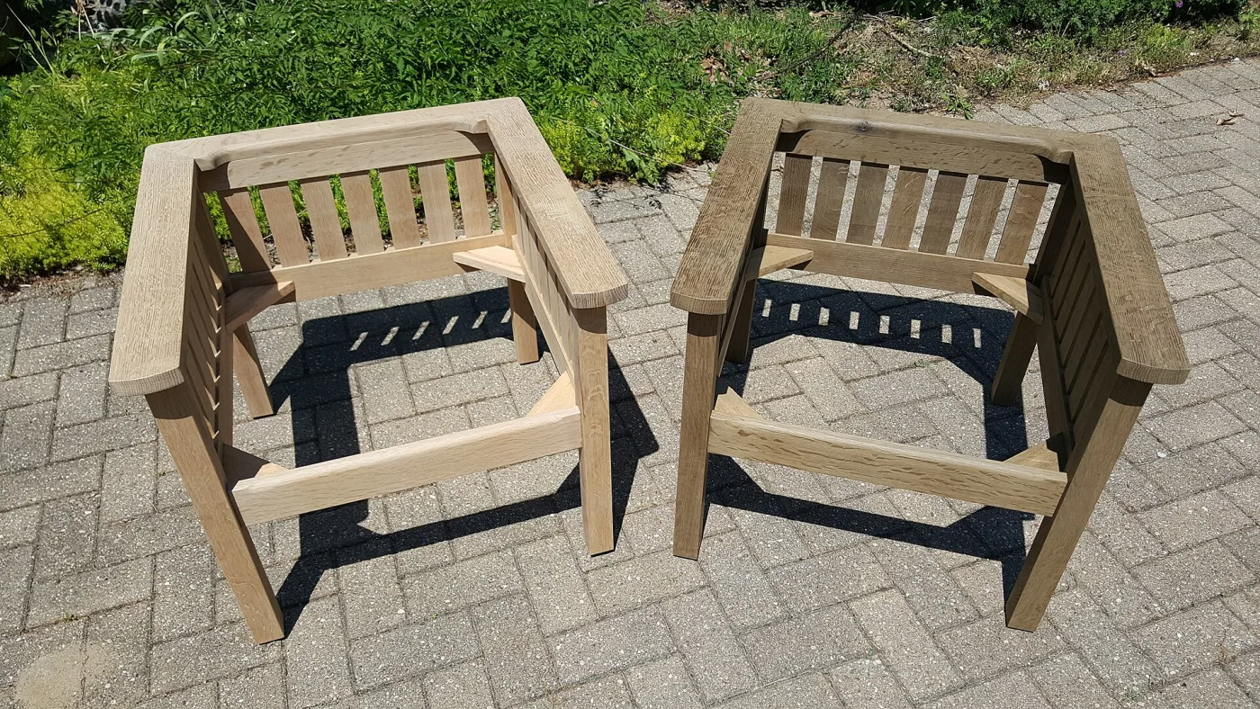 Two identical unfinished white oak chair frames with slatted sides standing side by side on a paved terrace in sunlight