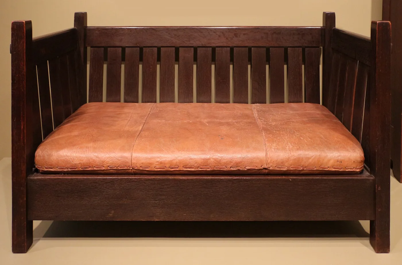 Low dark oak settle with vertical slats across the back and both ends, square corner posts, and a single long tan leather cushion laced along its front edge