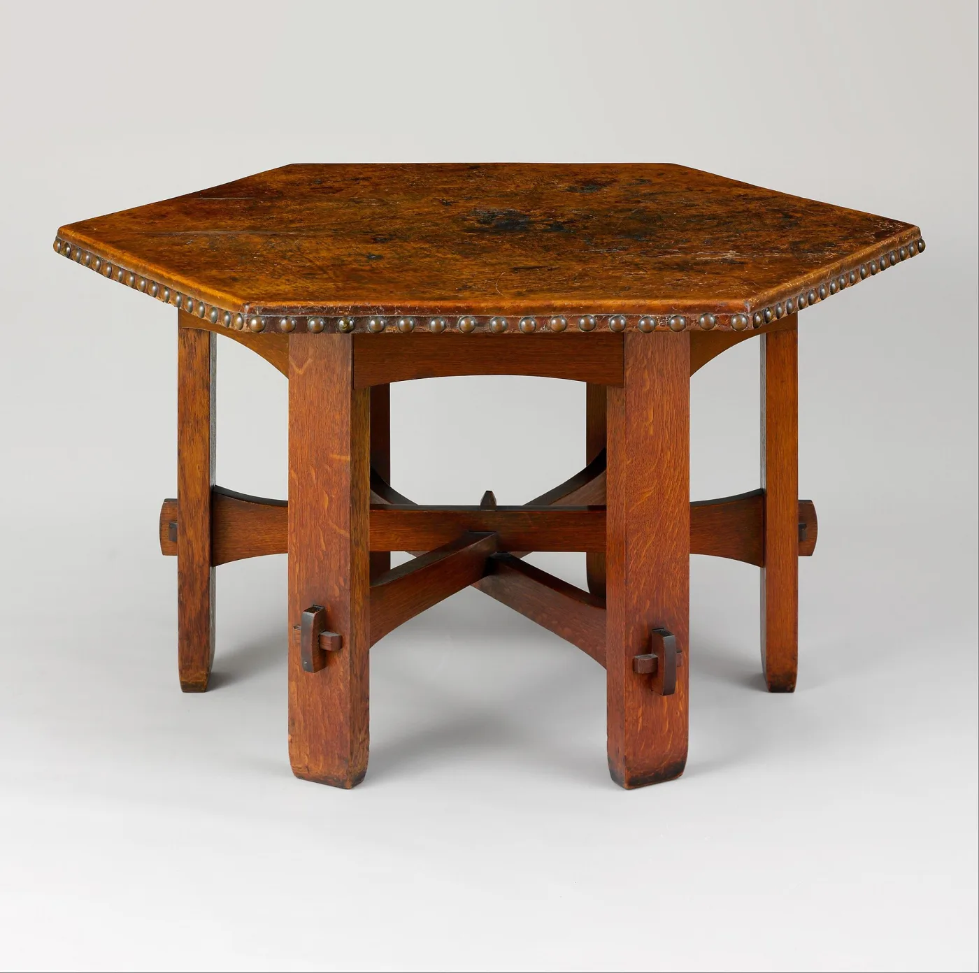 Hexagonal oak table with a leather top edged in domed nails, standing on splayed legs joined by cross stretchers locked with projecting wedge keys