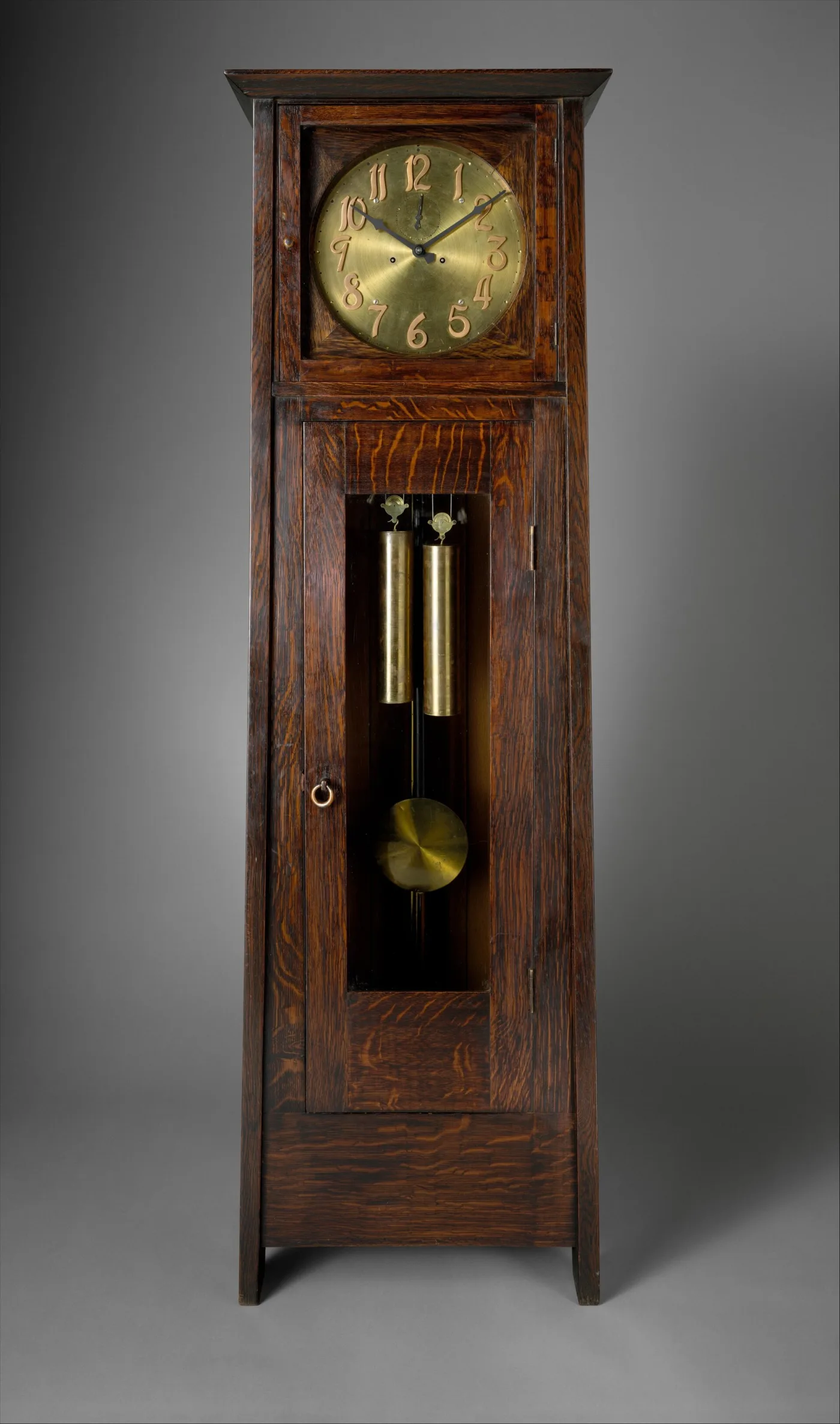 Tall oak clock case with a brass dial, a long glazed door showing weights and pendulum, and broad silvery flecks running across the grain of the stiles