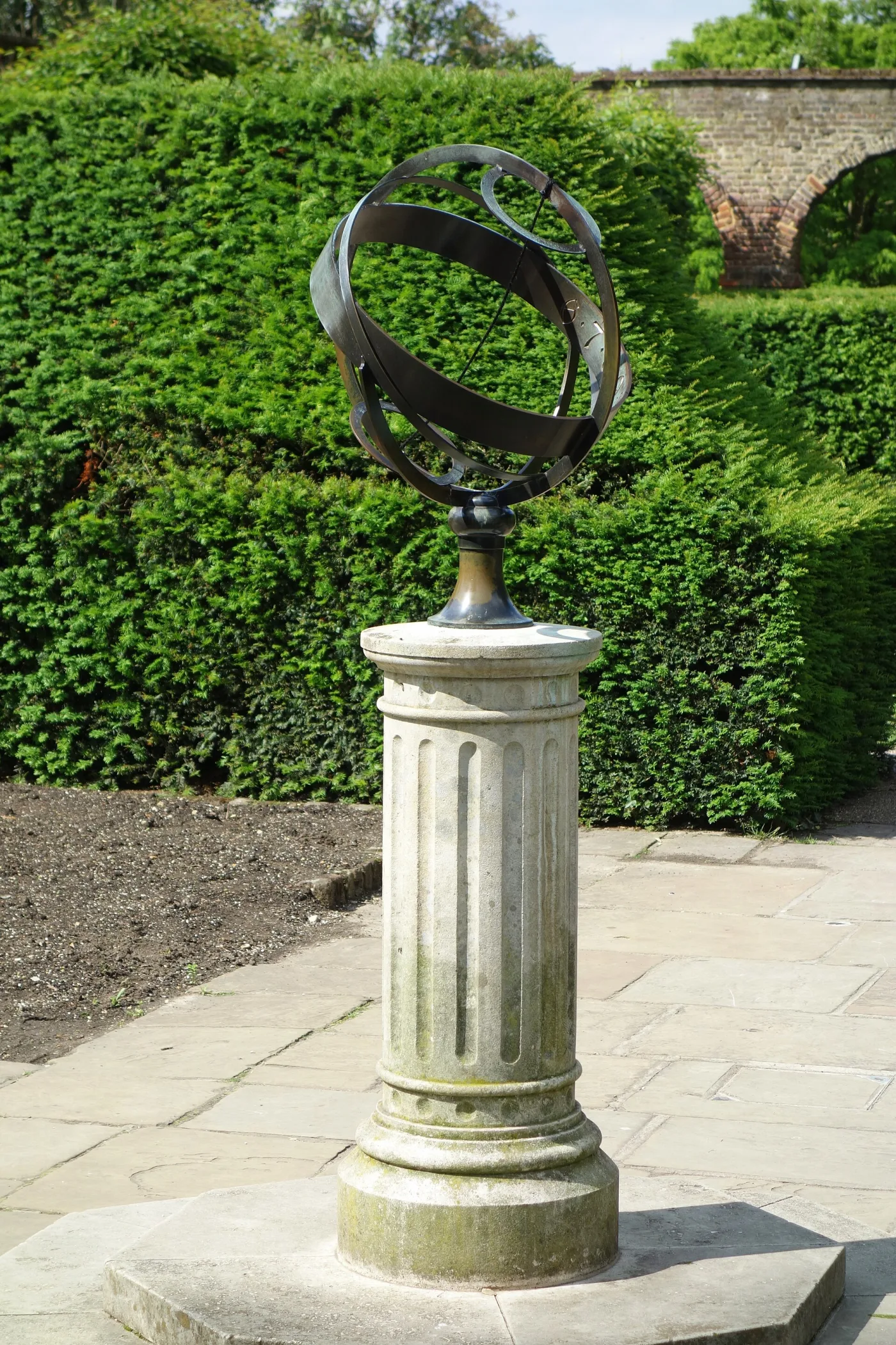 Dark bronze armillary sphere of broad flat rings on a fluted stone pedestal in a walled garden, with a rod through the polar axis