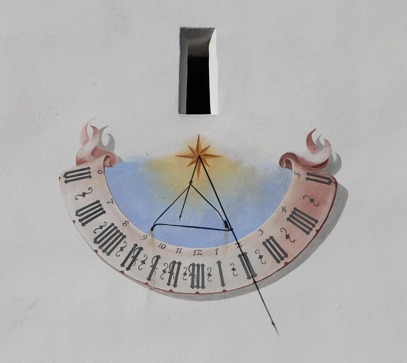 Painted semicircular wall sundial on the white render of a Tyrolean church, with blackletter Roman numerals on a pink scroll, a sunburst at the centre and an iron rod gnomon casting a shadow