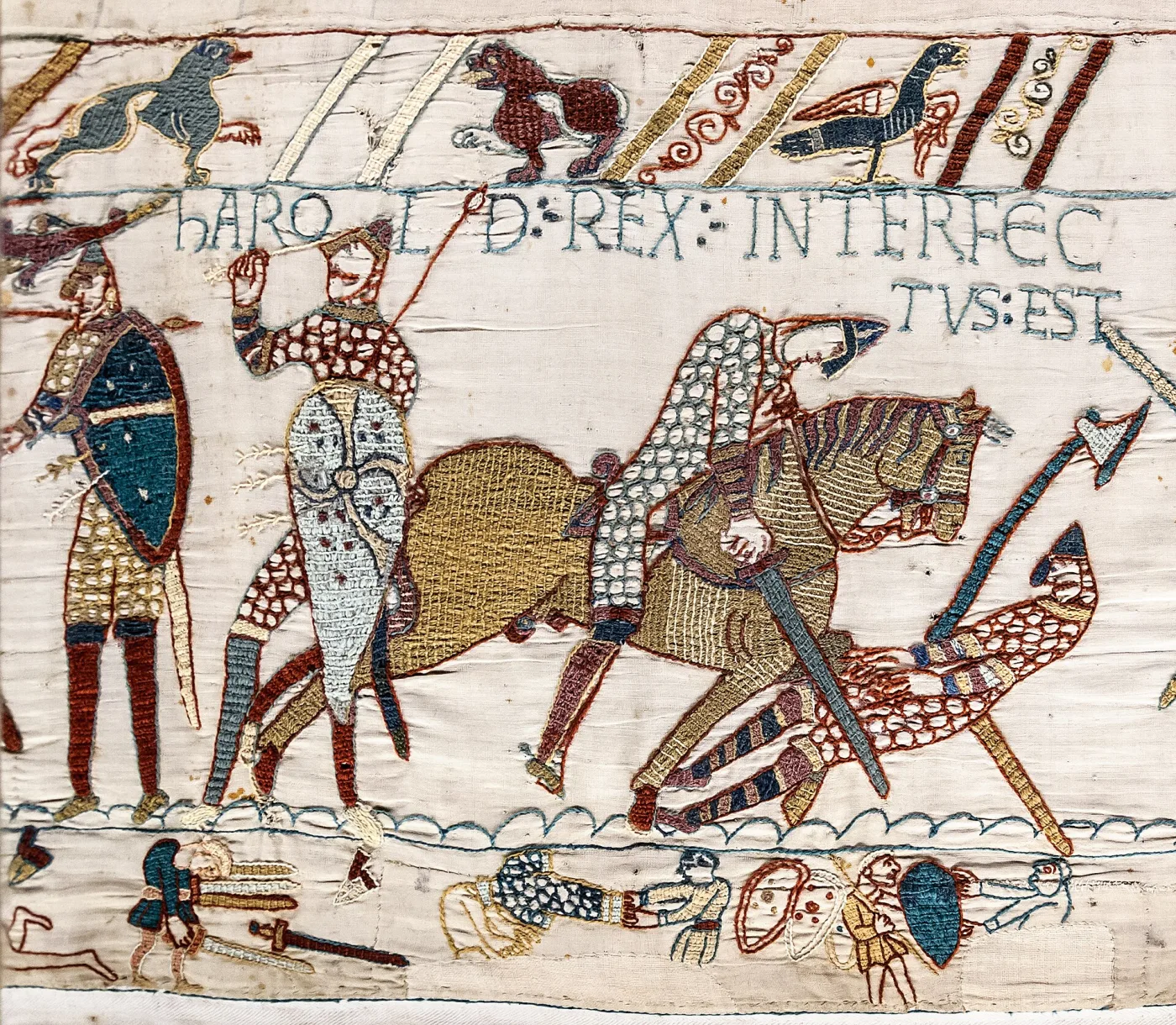 Scene from the Bayeux Tapestry showing mounted Norman knights and fallen figures in outlined wool stitching on plain linen, with the Latin inscription HIC HAROLD REX INTERFECTUS EST above