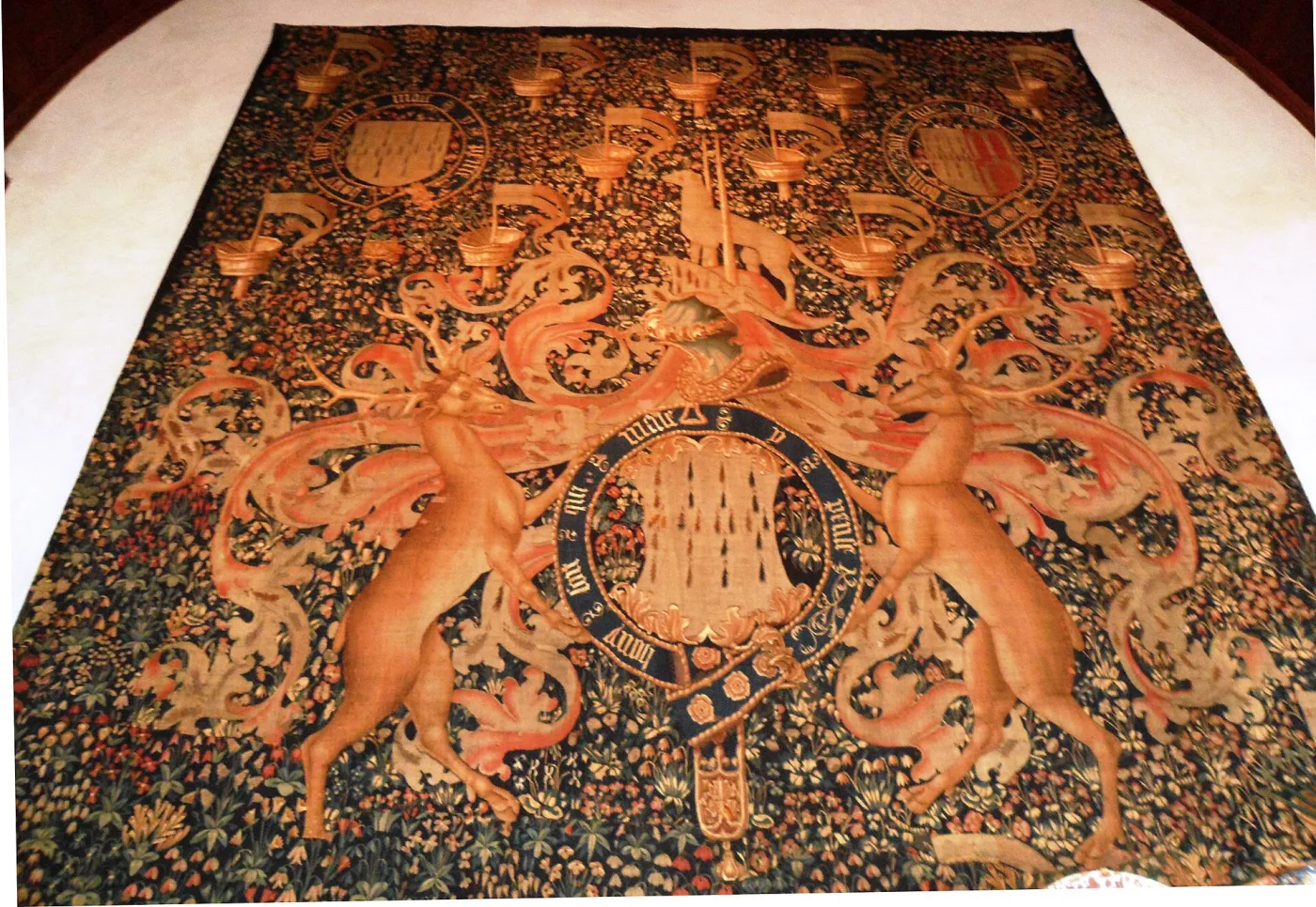 Millefleurs tapestry with a large central coat of arms and repeated heraldic badges scattered over a dense flowered ground, hung on a stone wall in a museum gallery
