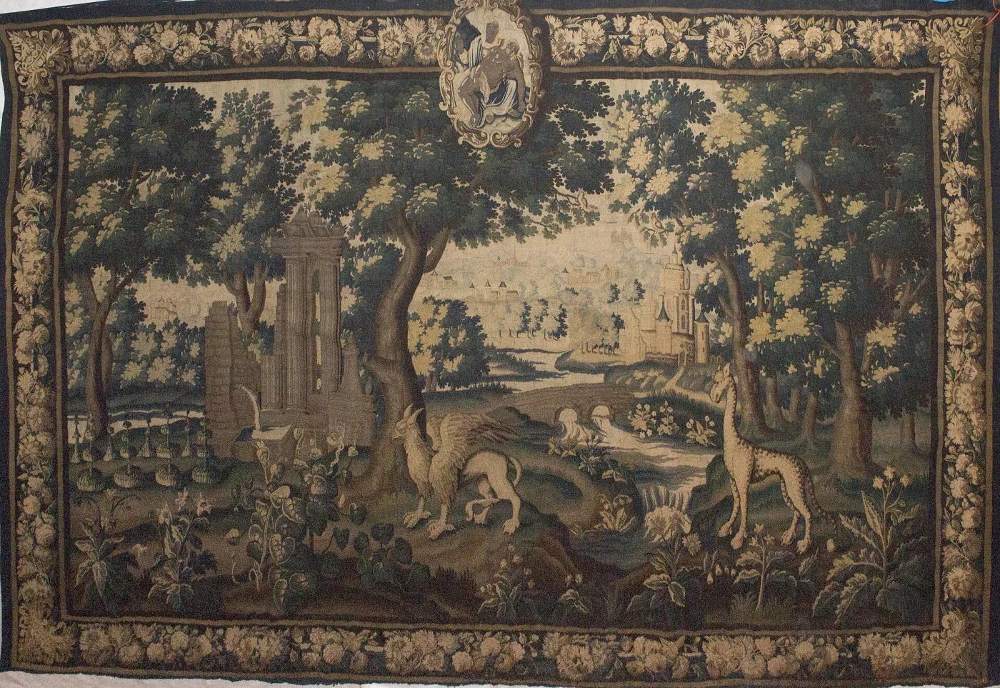 Aubusson verdure tapestry of about 1600 showing a wooded landscape in browns, blues and faded greens, with a spotted giraffe standing among trees on the right, a winged griffin lower left, and a walled town on a hill behind