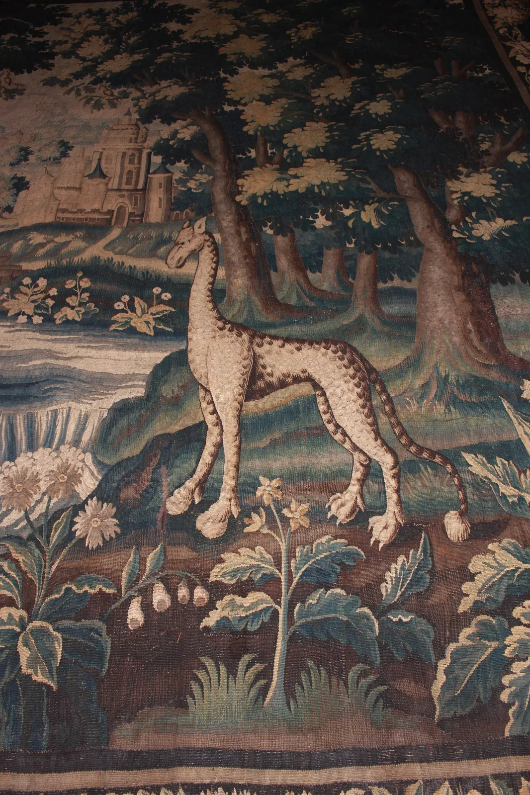 Close detail of a tapestry giraffe, its spotted coat rendered in interleaved bands of cream and brown wool, standing among stylised plants with a hilltop castle behind