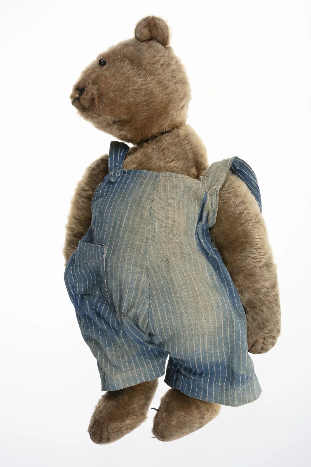 Beige-brown mohair teddy bear standing in blue-and-white striped cotton overalls, front view, with dark repaired paw pads and a worn muzzle