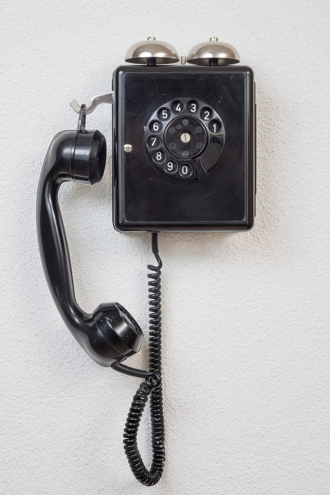 Black bakelite wall telephone with a rotary dial in the centre of the case and a handset hanging on a hook at the side, mounted on a plain white wall