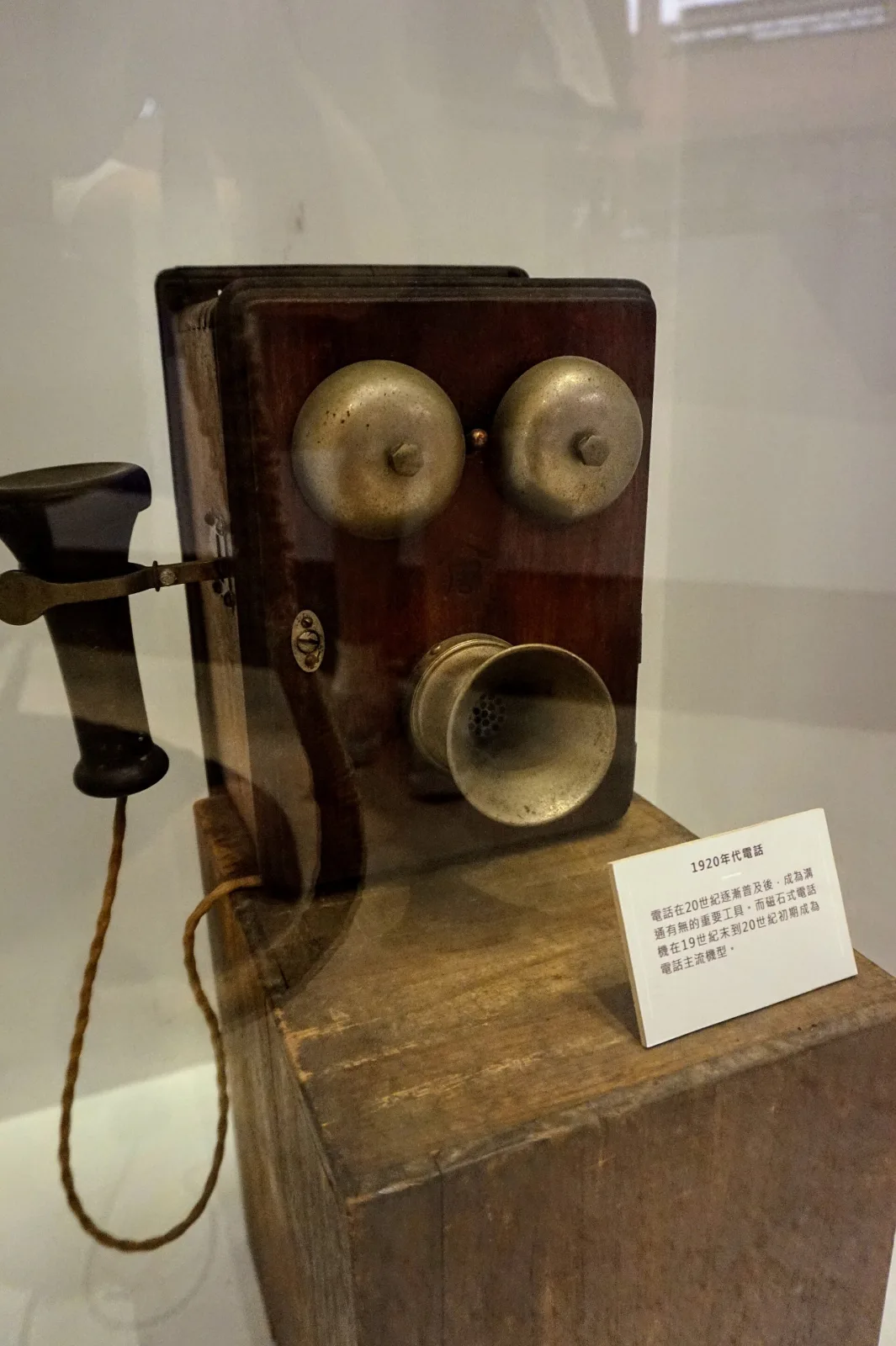Wooden wall telephone of the 1920s displayed in a museum case, with two brass bells, a projecting transmitter arm, a receiver hanging on a cord and an explanatory museum label beside it