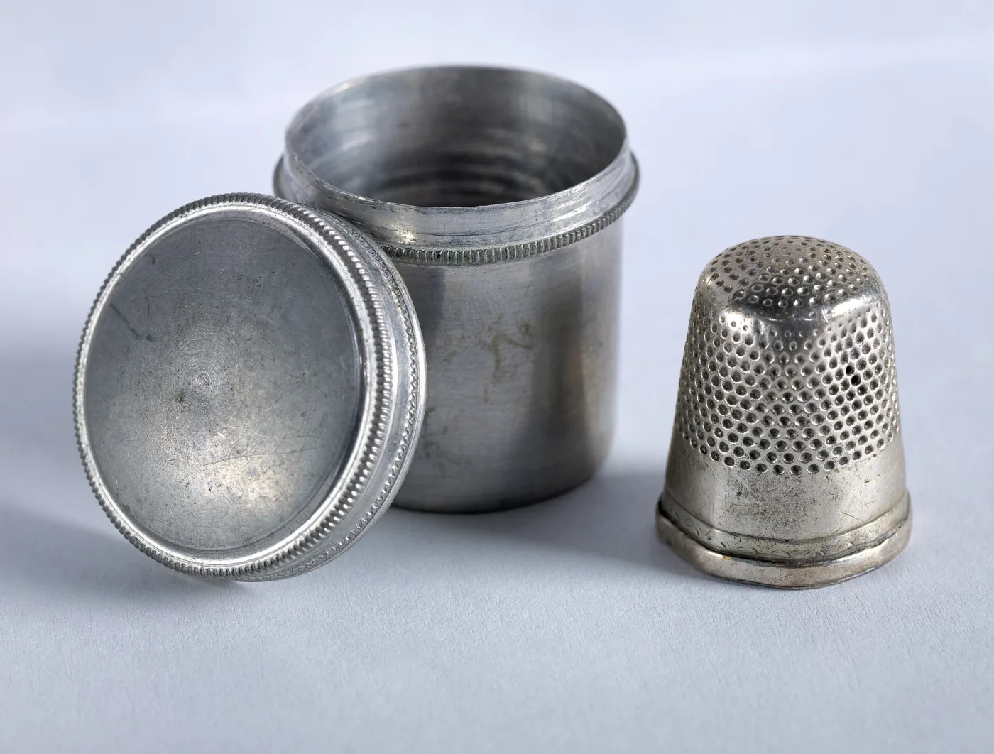 Silver thimble standing beside its cylindrical metal case, the case unscrewed into two halves lying open on a pale ground