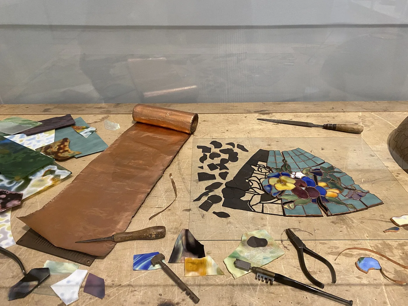 Workbench laid out with a paper pattern, sheets and offcuts of coloured glass, a partly assembled panel of cut pieces, glass cutters, pliers and a soldering iron