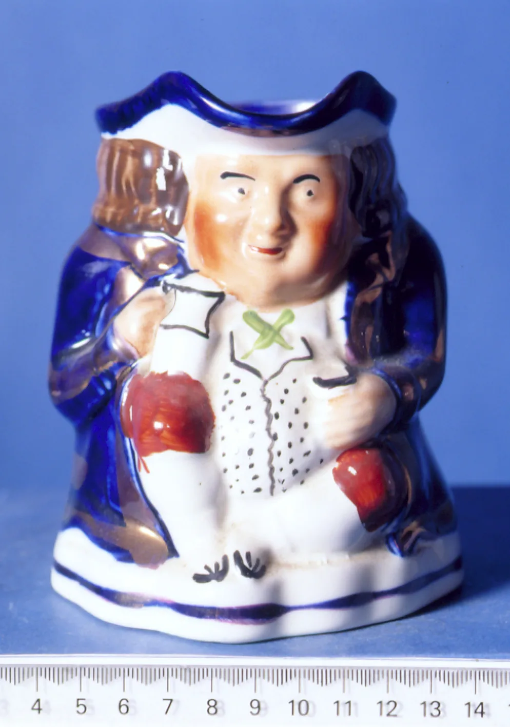 Full-length seated figure of a man in a blue coat and tricorn hat holding a jug, glazed in blue, white and flesh tones, photographed beside a centimetre rule