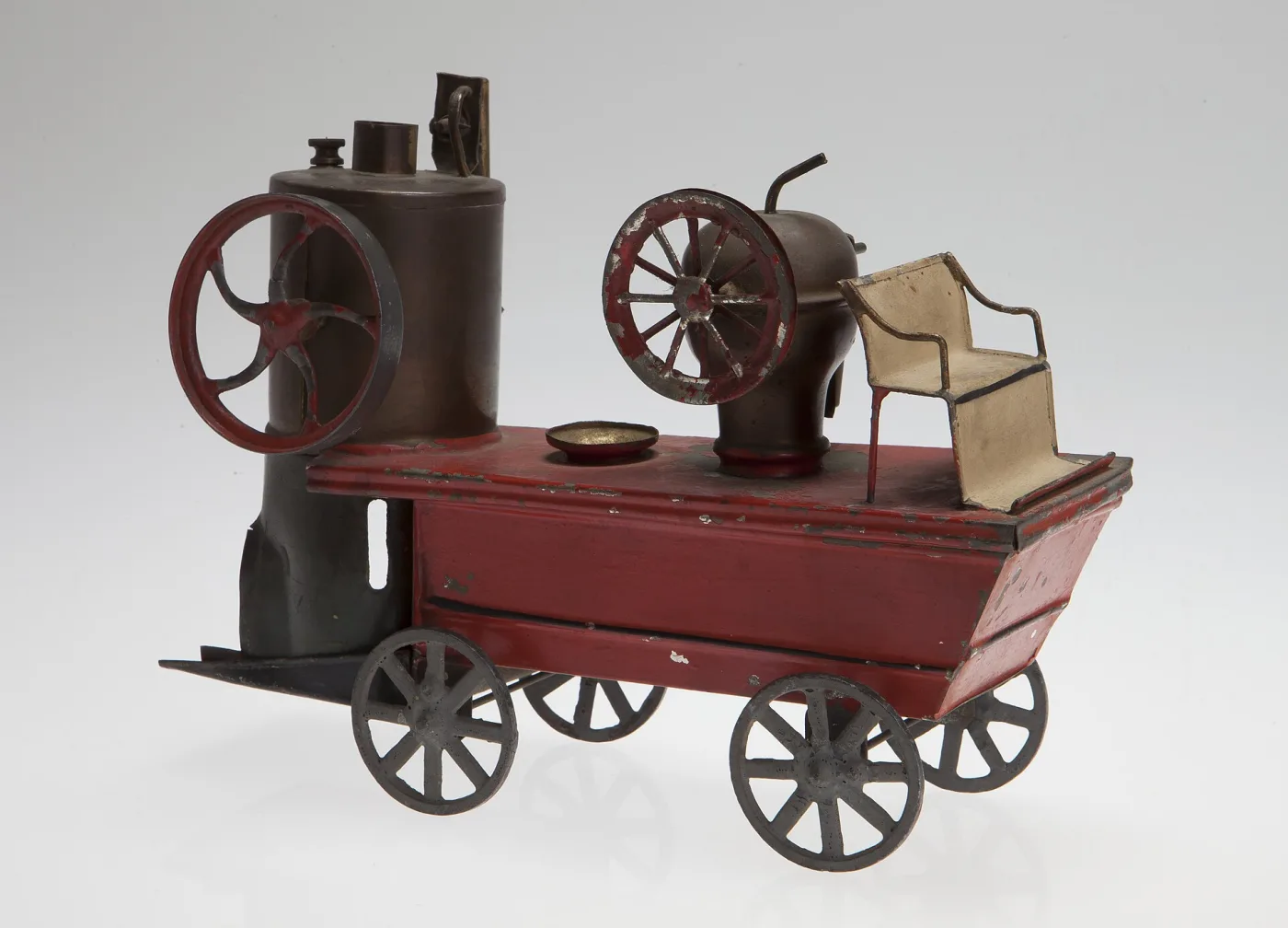 Nineteenth-century tin toy steam fire pumper painted dark red, with a tall black boiler, a large flywheel, spoked wheels and a hand-soldered platform, photographed against a plain grey ground