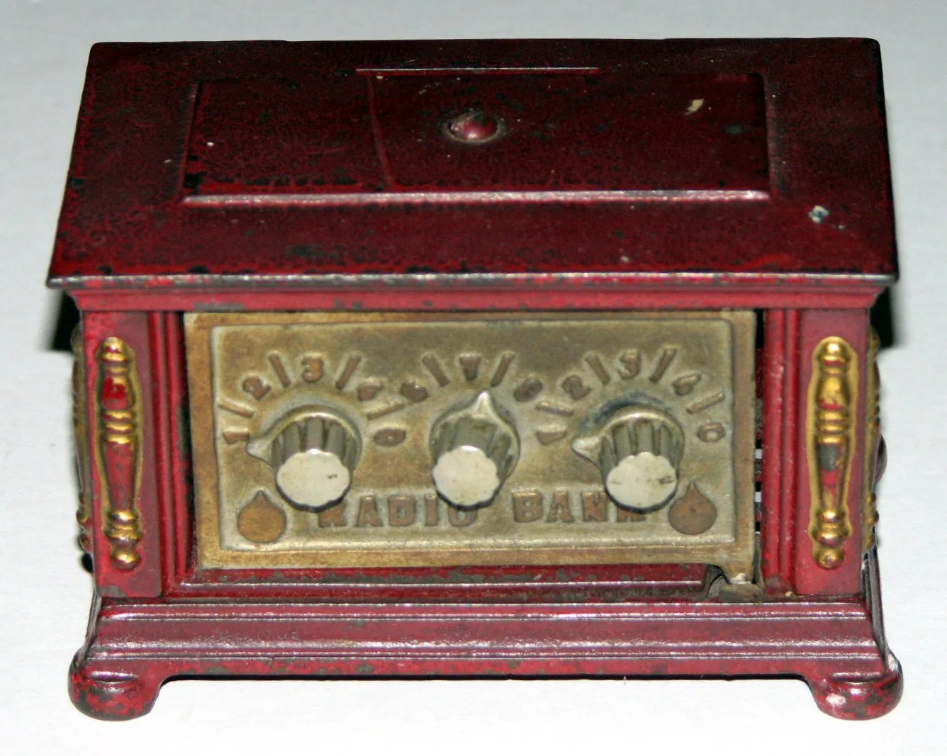 Cast iron toy bank in the shape of a radio cabinet, painted dark red with gilded dials and a combination lock on the front, standing on four small feet