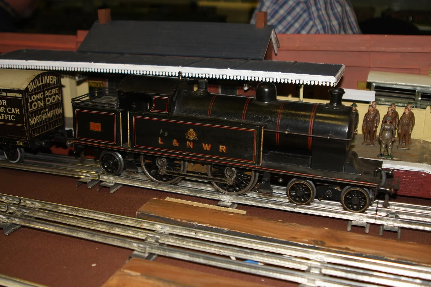 Large black tinplate tank locomotive lettered L and N W R with a crest, on tinplate track beside a station
