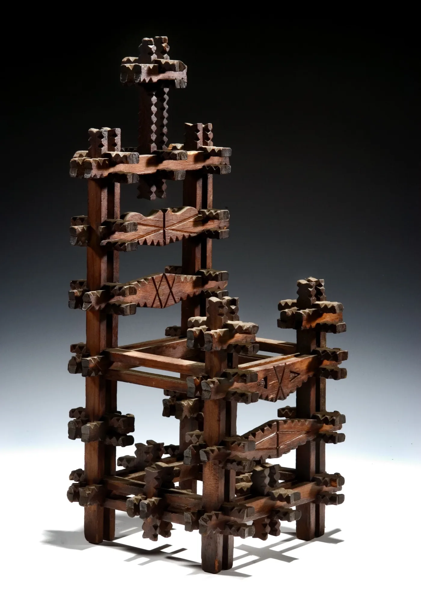 Open tower of notched wooden strips interlocked at right angles in stacked square tiers, with a cross rising from the top and light passing through the lattice