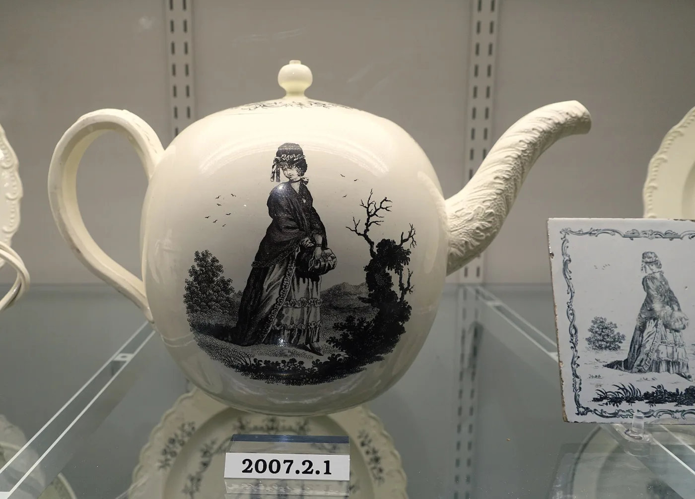 Cream-coloured globular punch pot printed in black with a standing woman in a hat and cloak in a landscape, displayed beside a framed print of the same figure