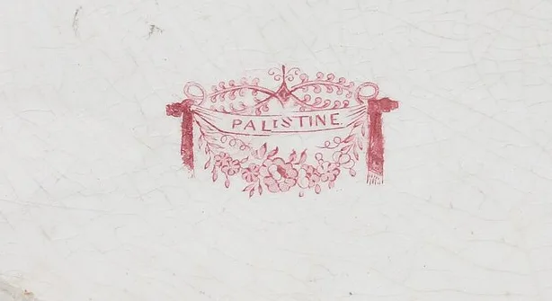 Close view of a printed pink cartouche of drapery and flowers on the crazed white underside of a platter, the pattern name Palestine lettered across a ribbon at its centre