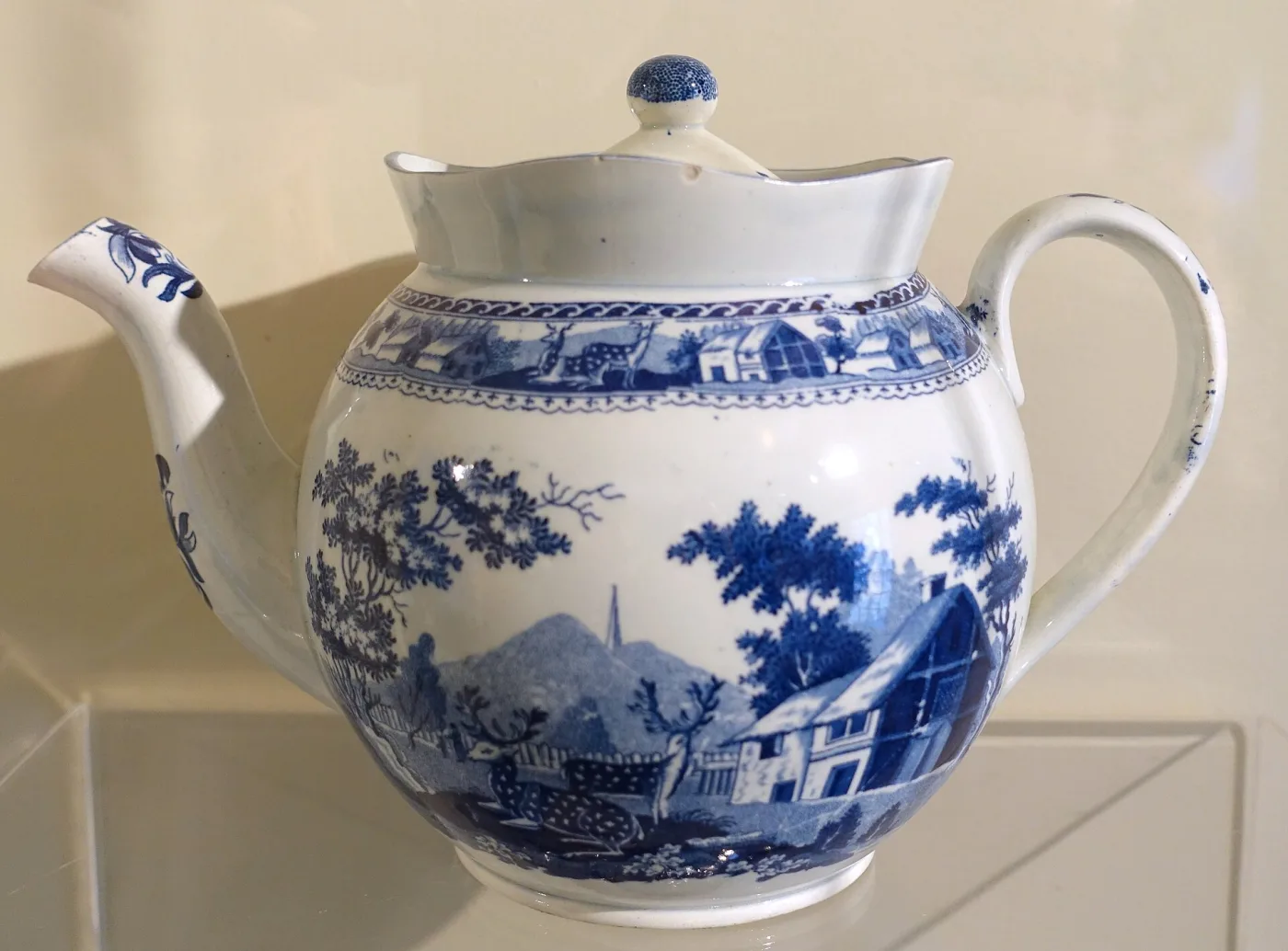 Globular earthenware teapot printed in blue with a landscape of buildings and trees, with a curved spout, loop handle and domed lid