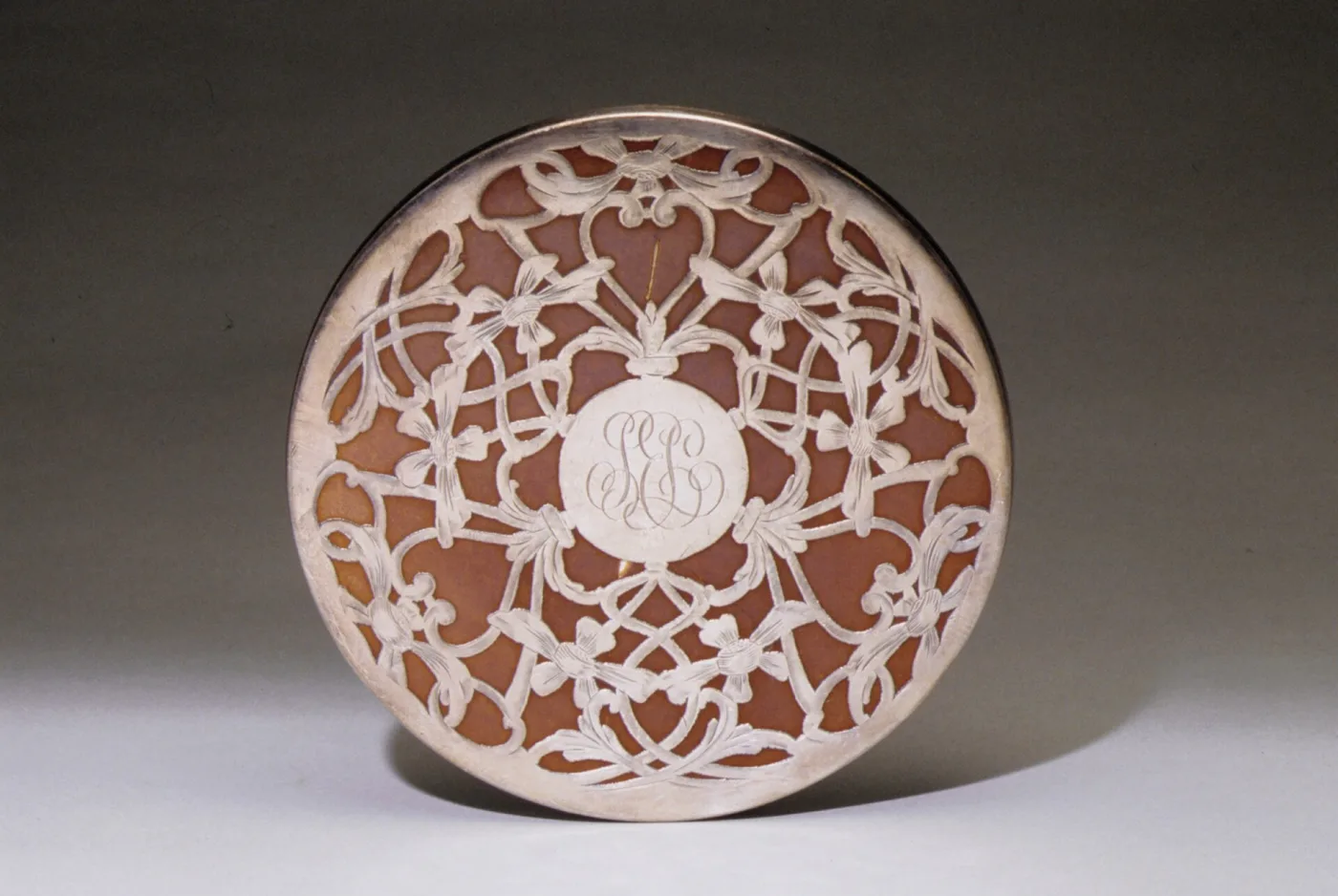 Round trivet with a warm brown ceramic body overlaid with pierced and engraved silver scrollwork and a script monogram at the centre