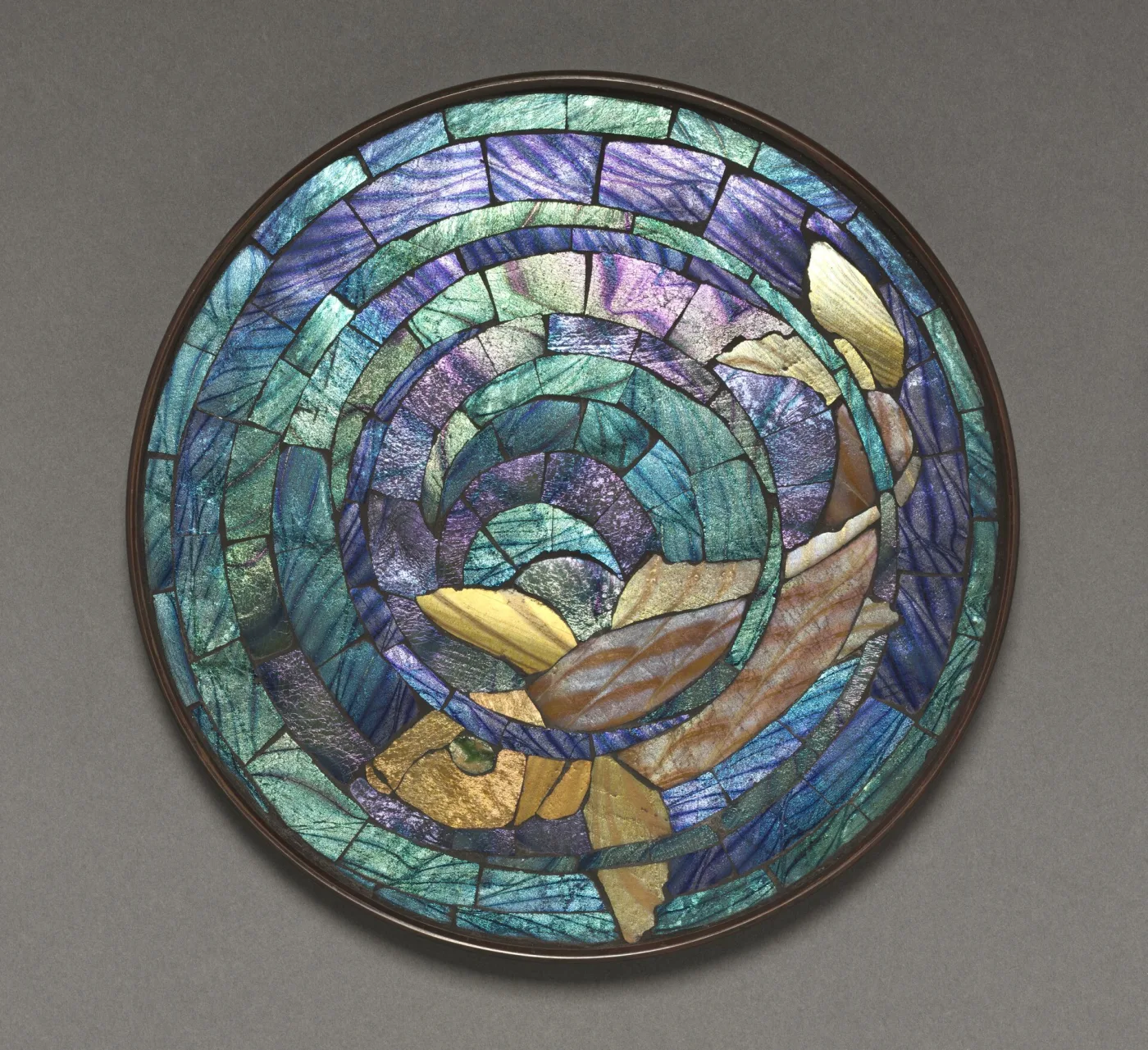 Round trivet with a bronze rim, its whole top filled with iridescent blue, green and gold glass mosaic laid in concentric bands