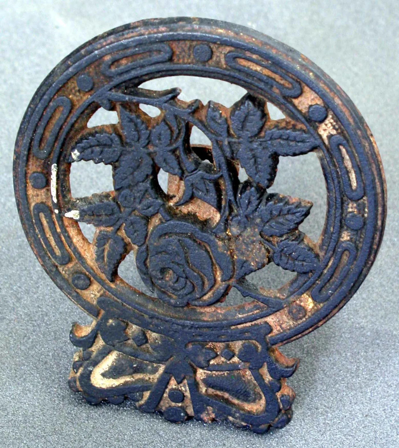 Circular cast iron pot trivet with a rose and leaves in relief inside a looped border, its black coating worn away to bare metal on the raised edges