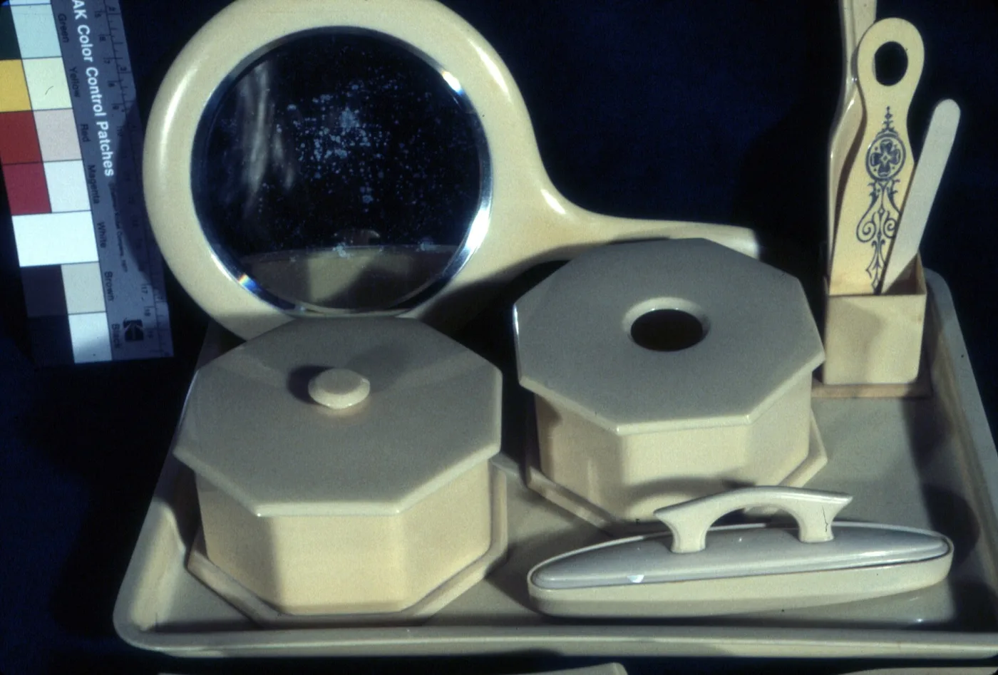 Ivory-coloured celluloid dresser set on a matching tray: a round hand mirror, two octagonal covered boxes, one lid pierced by a round hole, a clothes brush and a buffer in a stand, beside a photographic colour reference card