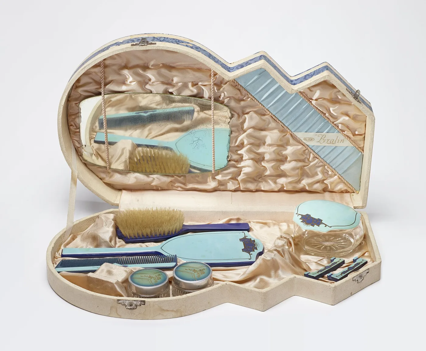 Shaped presentation box lined in peach satin holding a pale blue and cobalt plastic hairbrush, comb, hand mirror, glass jars and nail tools, with a printed DU PONT Pyralin label sewn into the lid lining