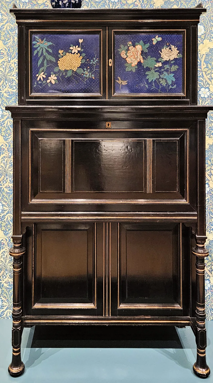 Tall black cabinet on turned legs, its upper doors set with two blue enamel panels of flowers, the lower case in plain ebonised panels with fine gilt lines