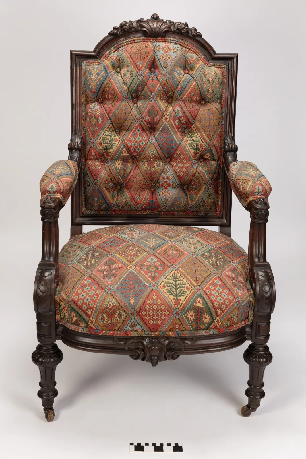 Dark walnut armchair photographed square-on against a plain ground with a scale bar, its back and seat deeply buttoned in a red and blue geometric fabric, on turned and fluted legs with casters