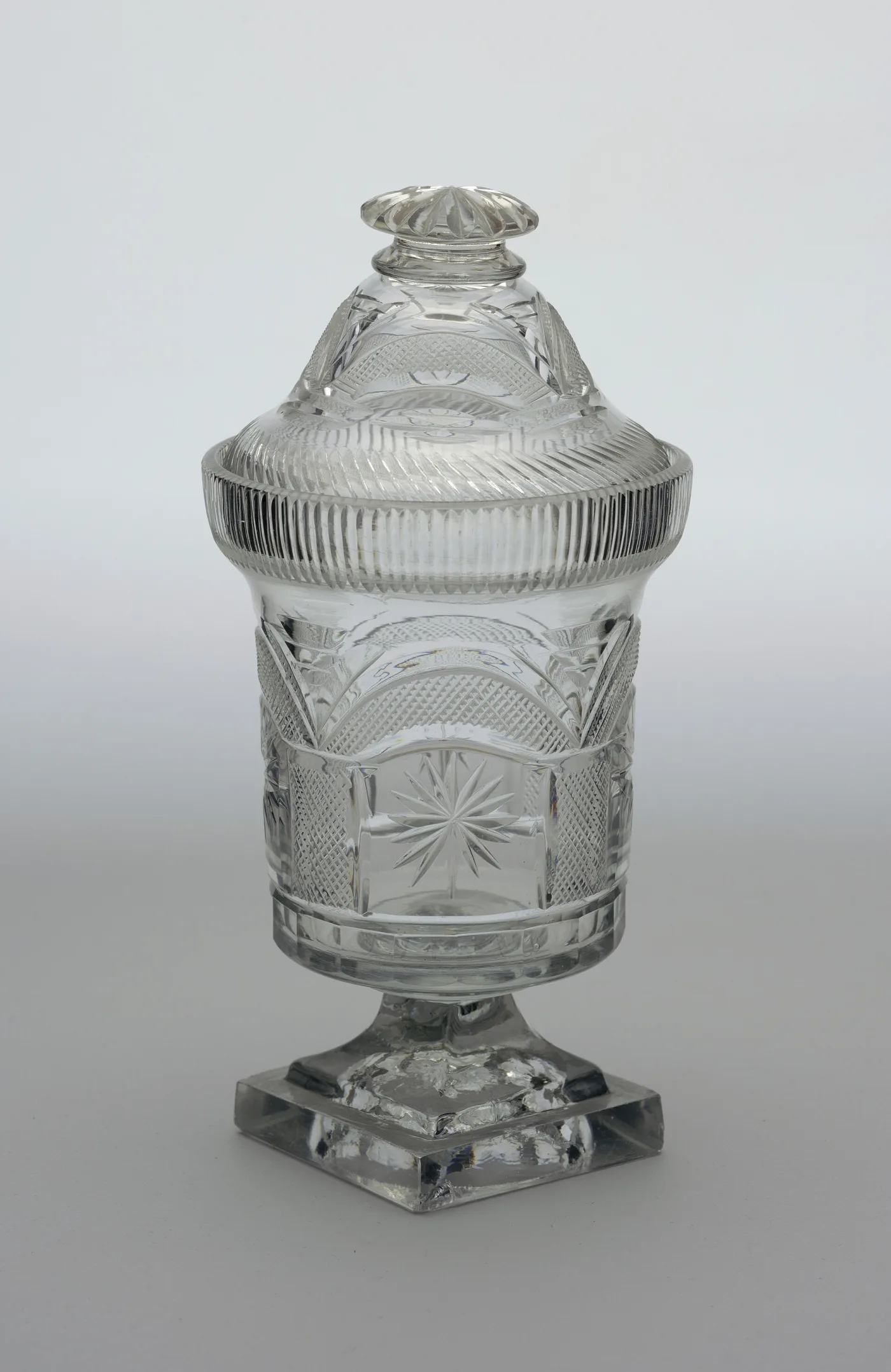 Covered crystal jar on a square pedestal foot, cut with arched panels of fine diamonds enclosing eight-pointed stars, under a domed and fluted lid