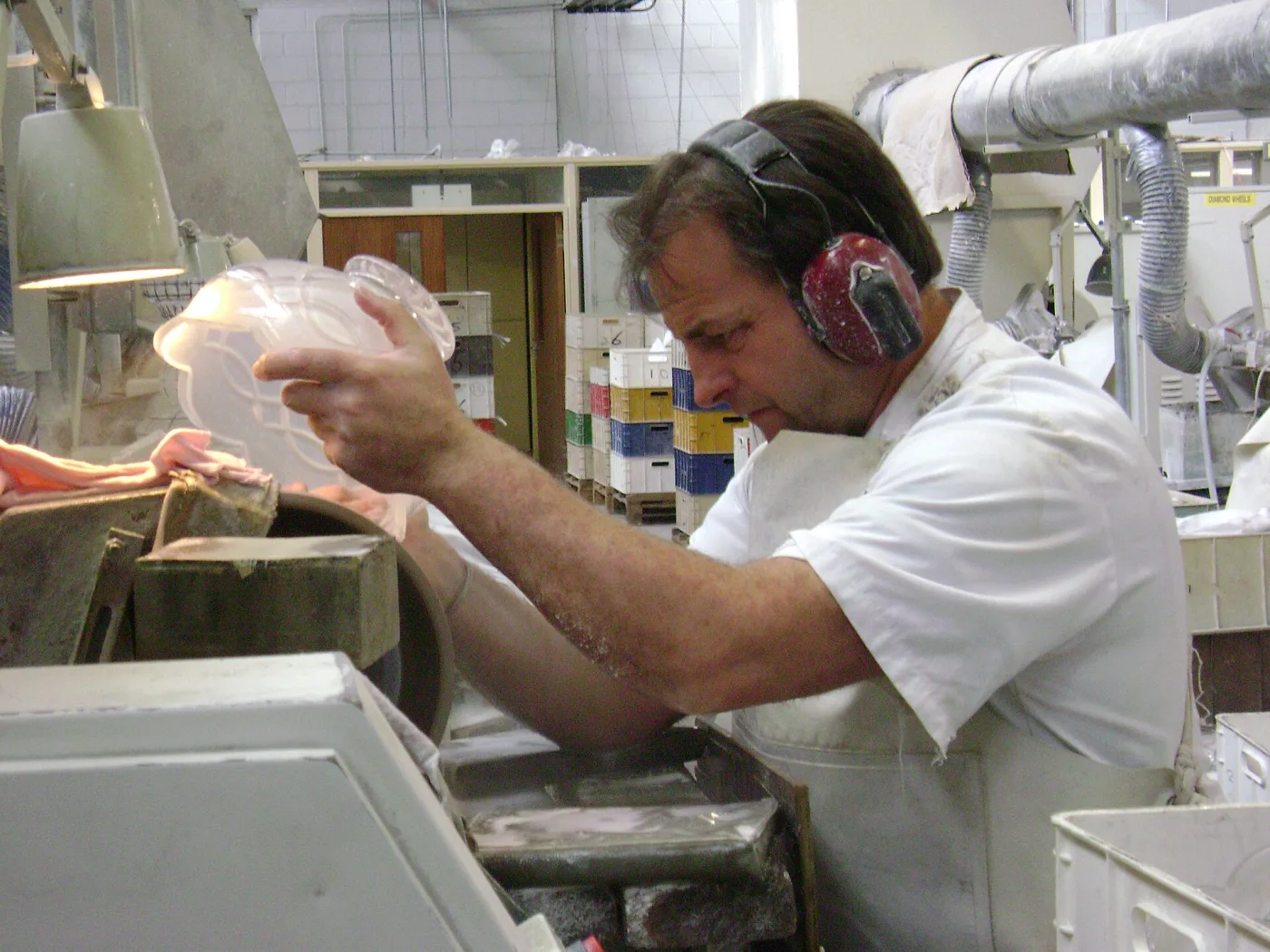 A cutter in ear defenders at the Waterford works holding a crystal dish against a rotating wheel, the surface white with grinding slurry