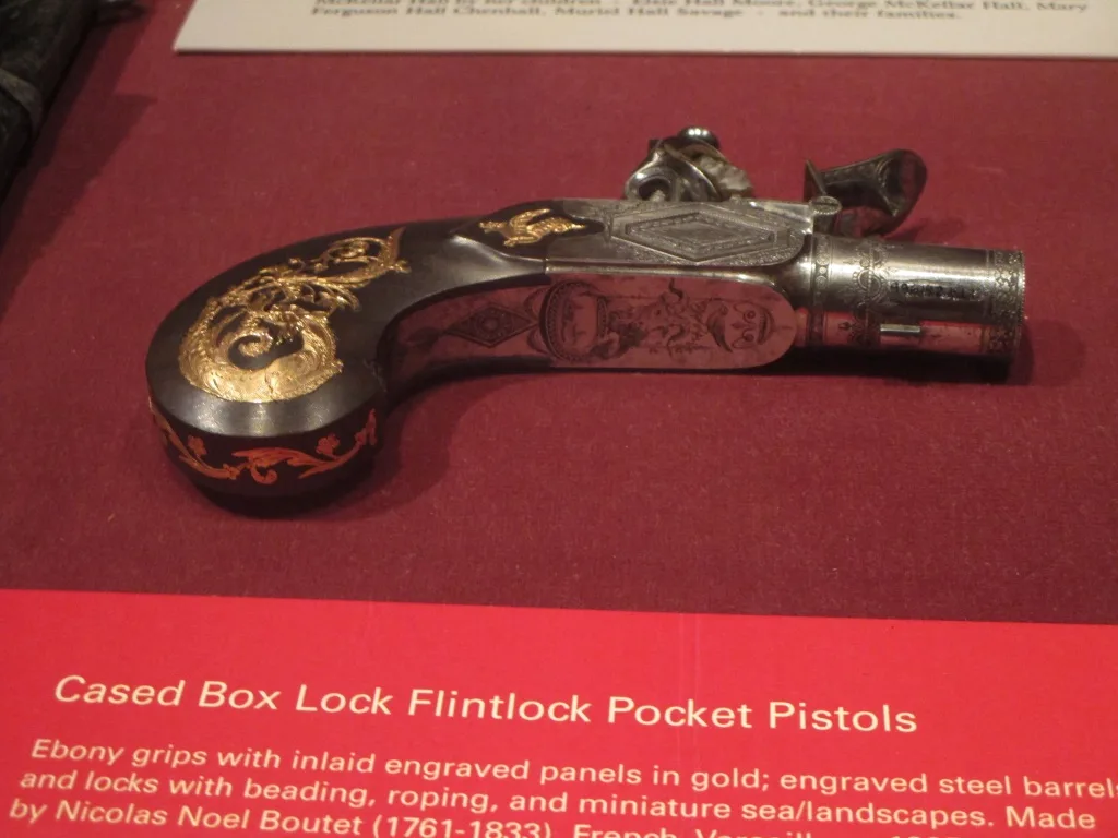 Small flintlock pocket pistol with a dark ebony grip inlaid with engraved gold panels and a short engraved steel barrel, displayed on a red mount beside its printed museum label