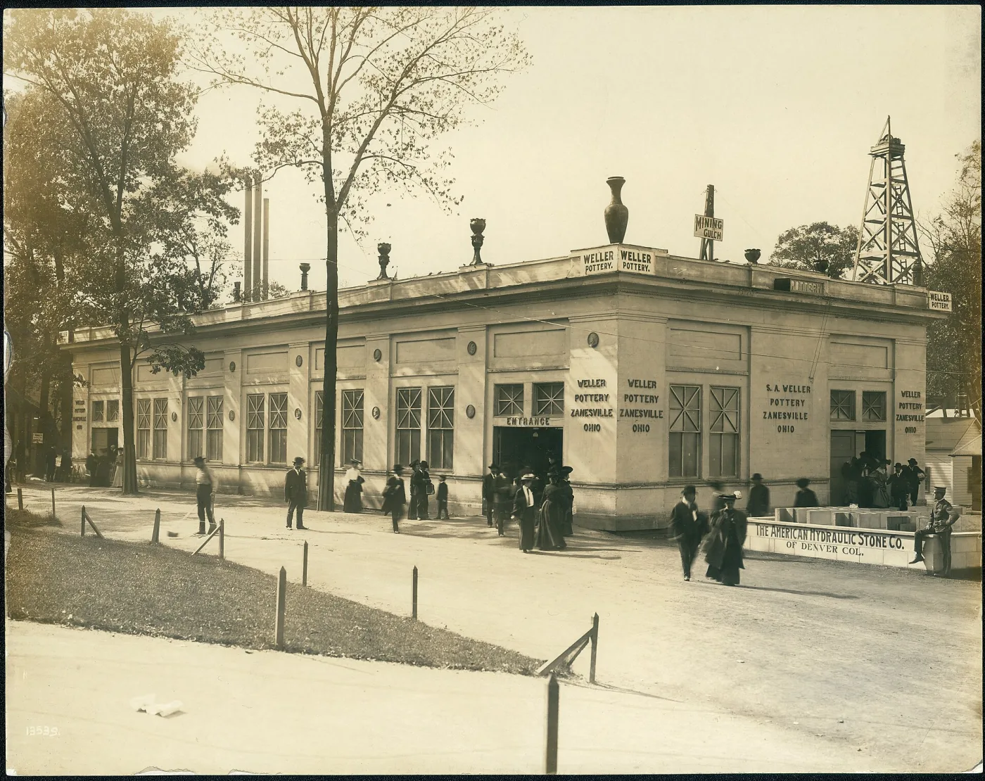 Sepia photograph of a single-storey stone exhibition building with a row of large vases standing along its roofline, people walking past on an unpaved street with a wooden tower behind