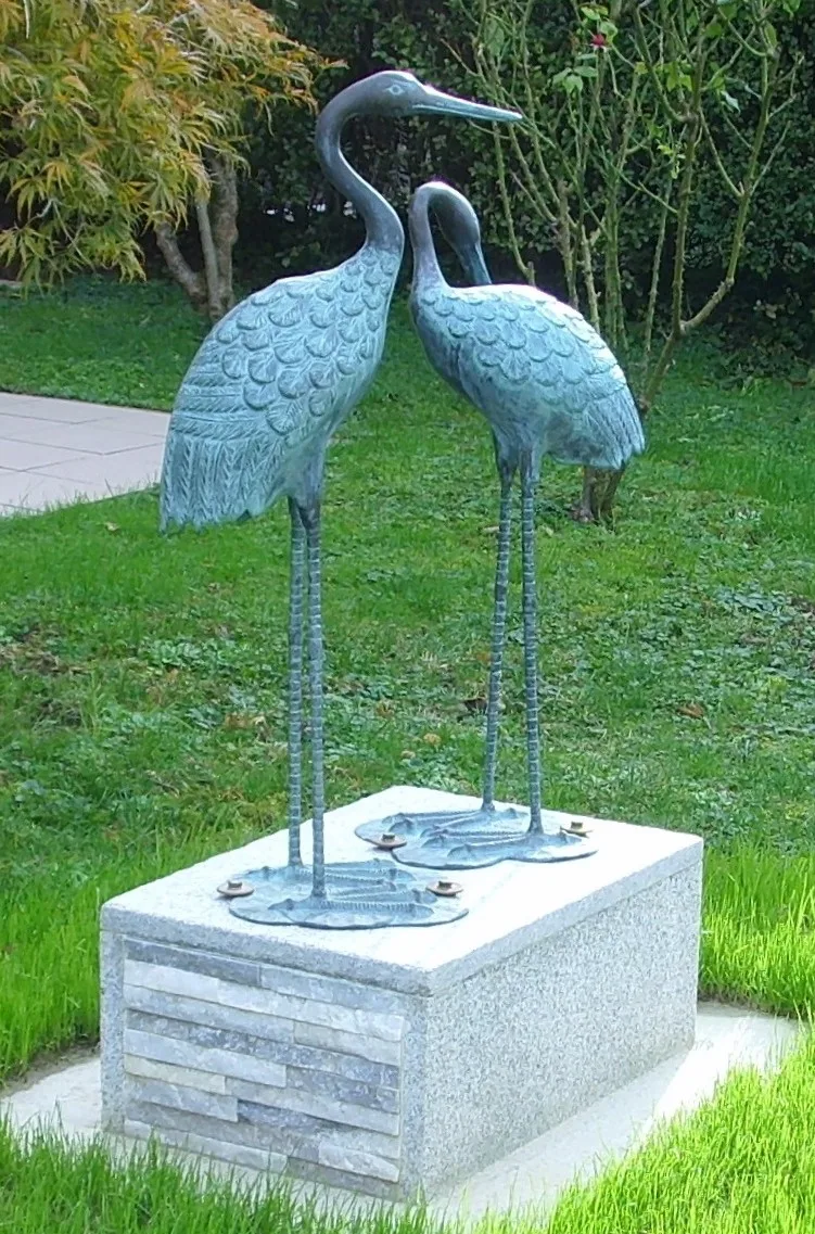 Two life-size bronze cranes with long legs and raised beaks, their surfaces an even pale blue-green, mounted on a pale stone plinth on a lawn