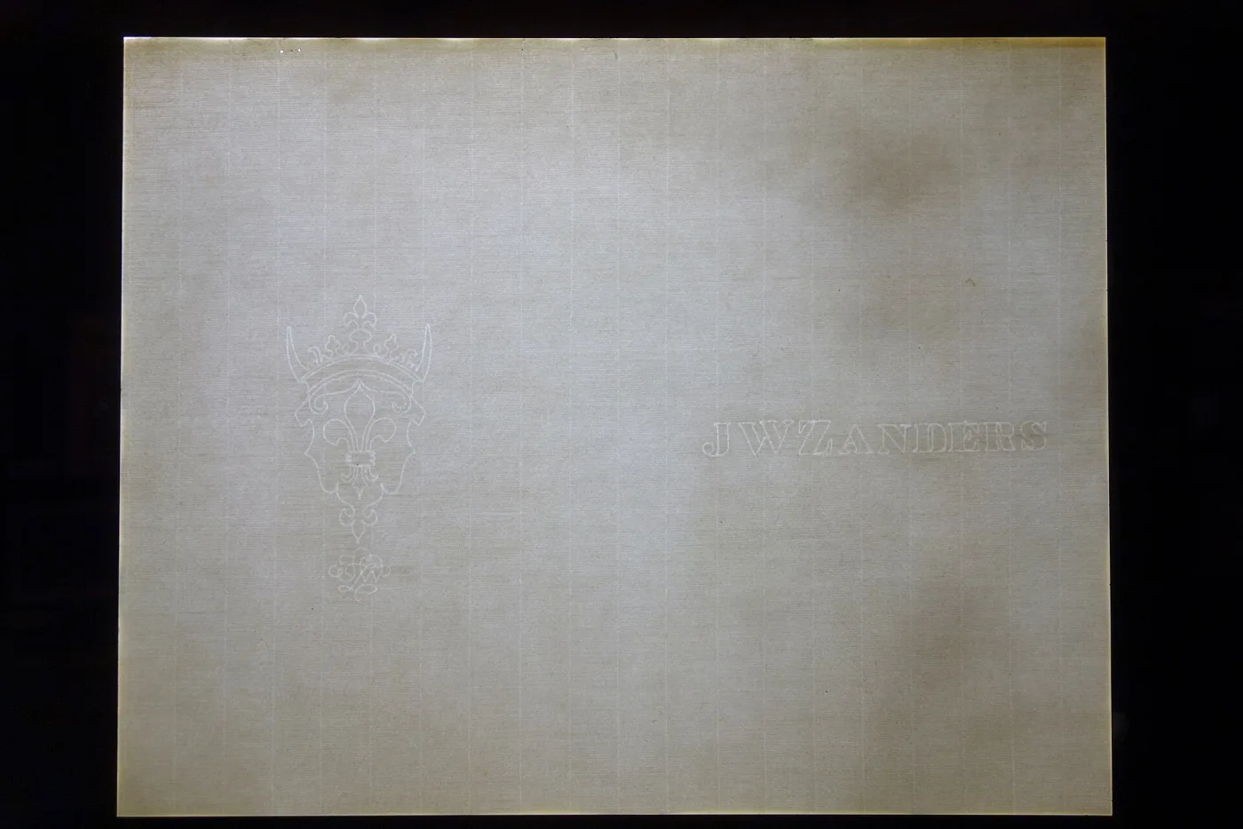 A sheet of pale laid paper photographed against a dark ground with light passing through it, showing fine horizontal lines, widely spaced vertical lines, a crowned fleur-de-lis watermark at the left and the letters J W ZANDERS at the right