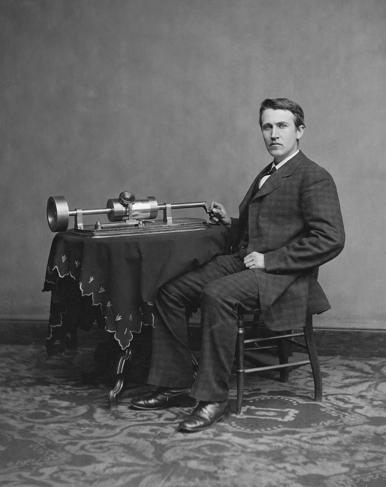 Studio photograph of about 1878 showing a seated young Thomas Edison beside a table carrying his tinfoil phonograph, a cylinder machine with a large crank handle