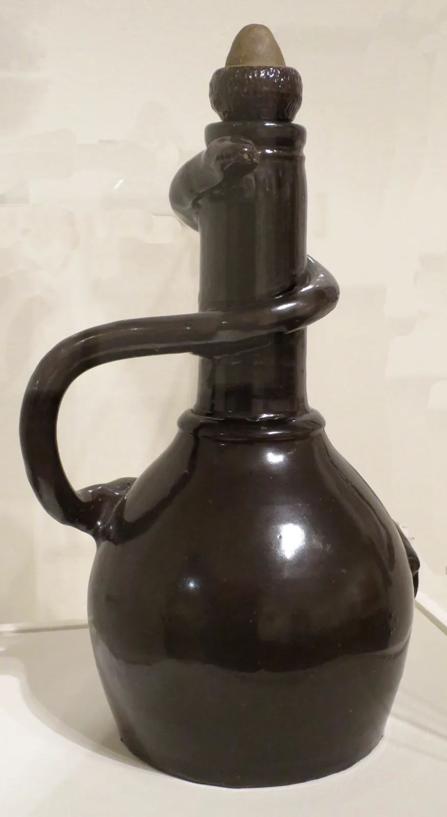 Tall dark brown glazed stoneware jug with a modelled snake coiled twice around its narrow neck, its head raised at the shoulder, a strap handle at the side and a stopper in the mouth