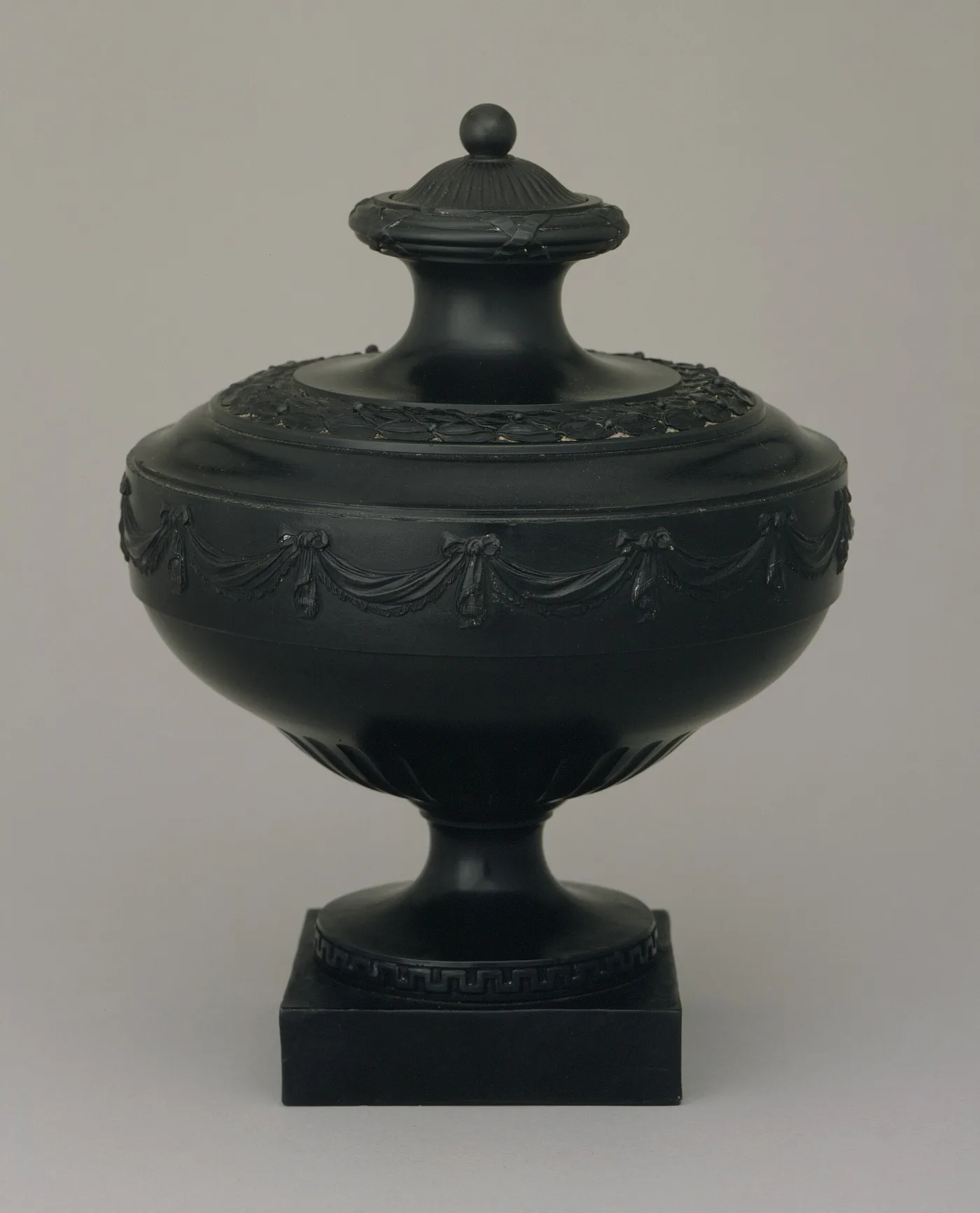 Neoclassical black basalt covered vase on a square plinth with swagged drapery ornament in the same black body